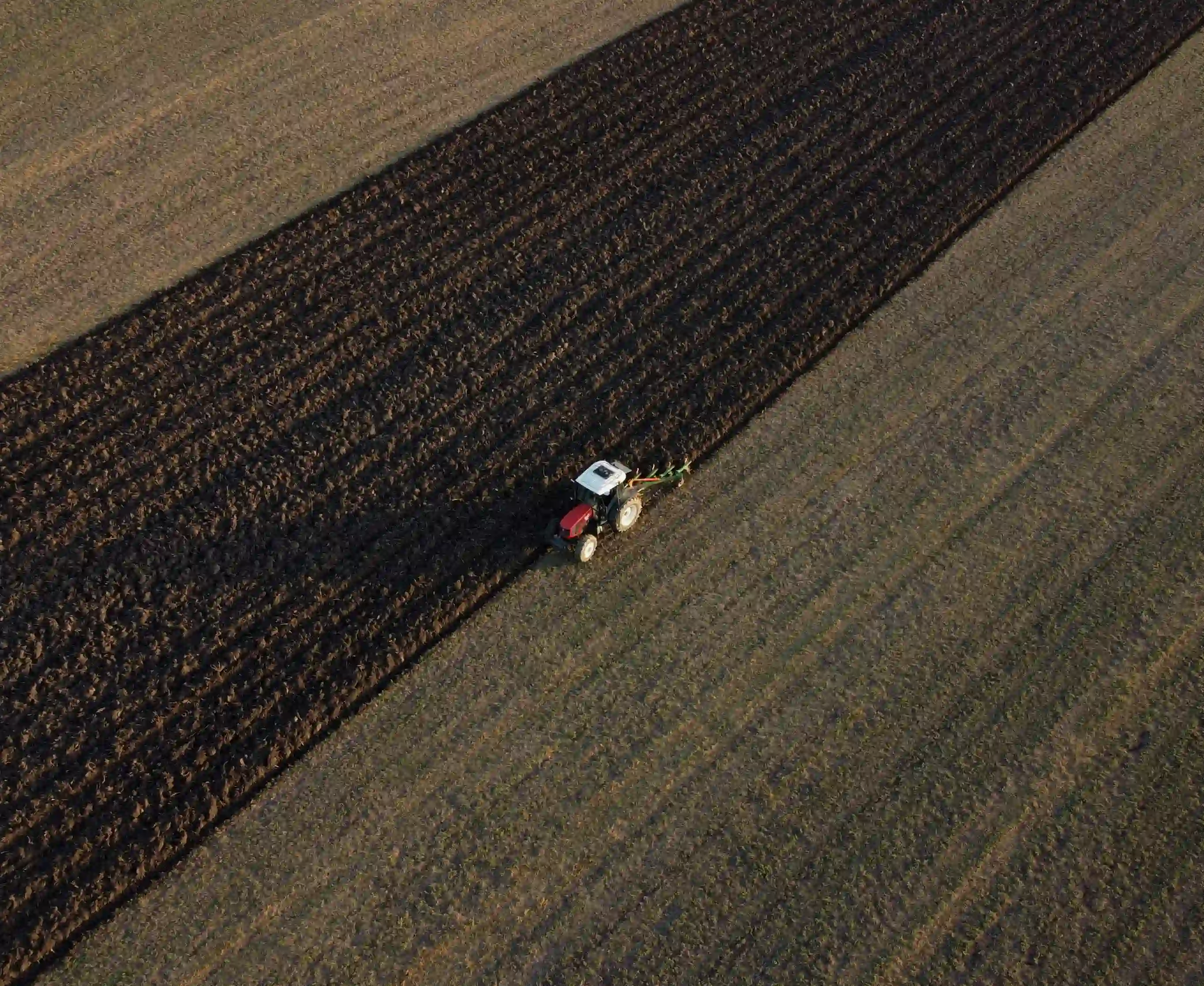 Aerial view of a tractor plowing a massive field, turning over dark soil in stark diagonal lines.