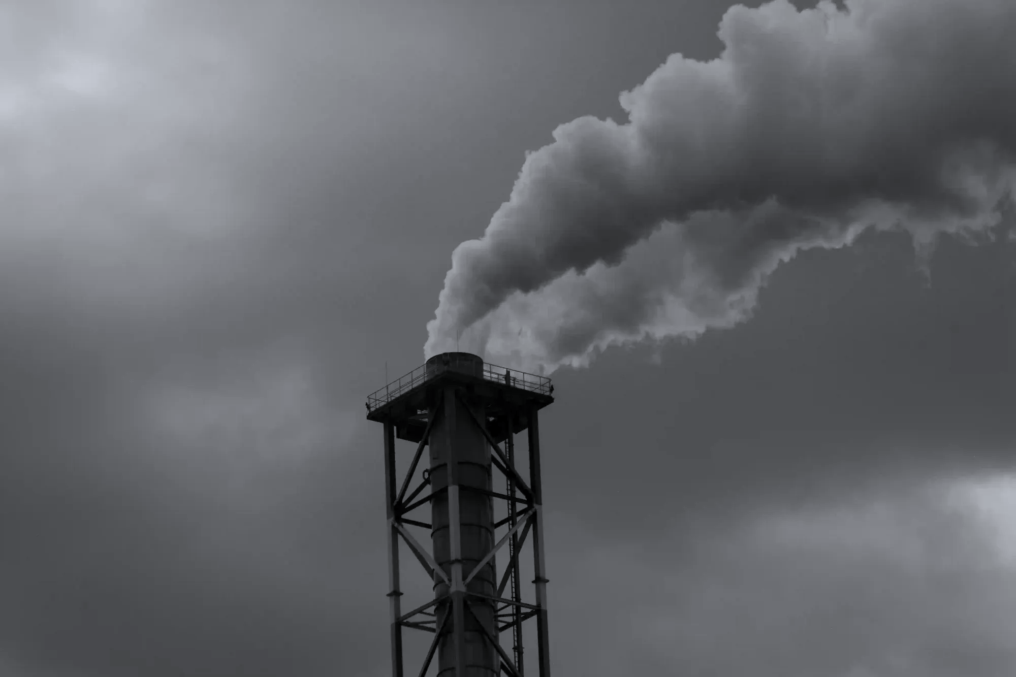 A tall industrial chimney emitting a thick plume of smoke against a moody, overcast sky in black and white, representing heavy industry.