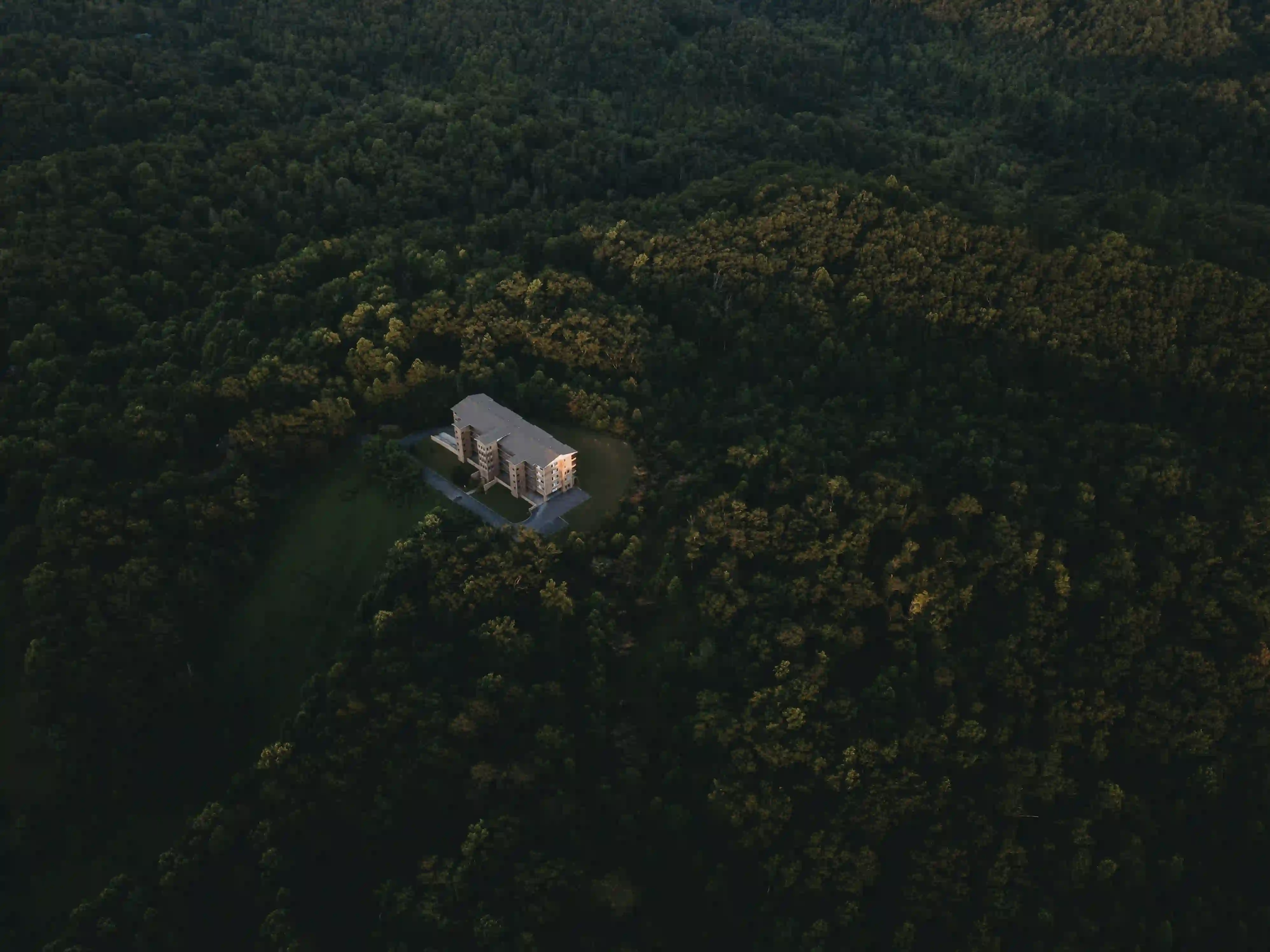 Aerial view of a solitary geometric building completely surrounded by a vast dark forest, symbolizing human isolation from nature.