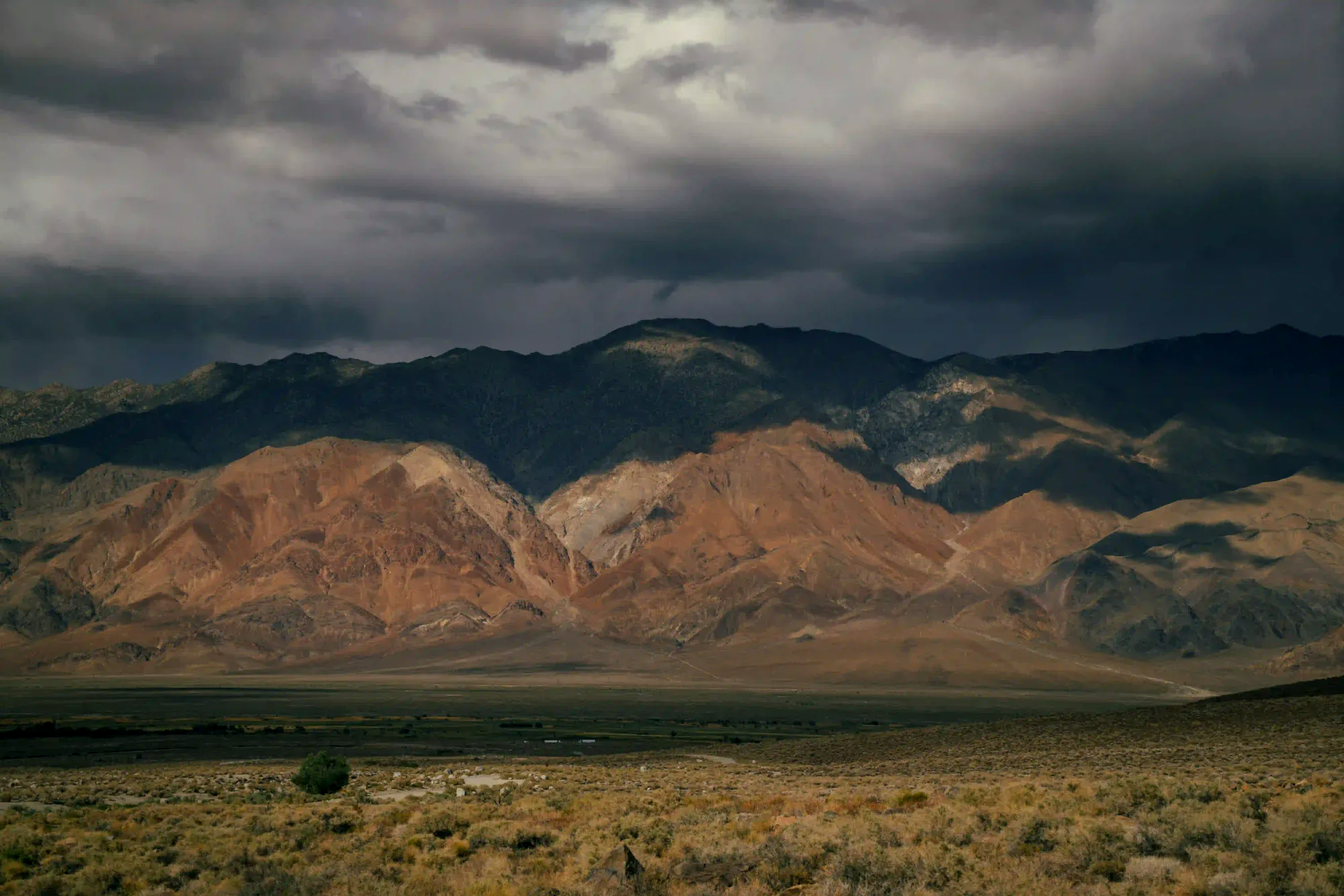 A vast, dry mountainous landscape under heavy, dark storm clouds, with dramatic sunlight hitting the peaks.