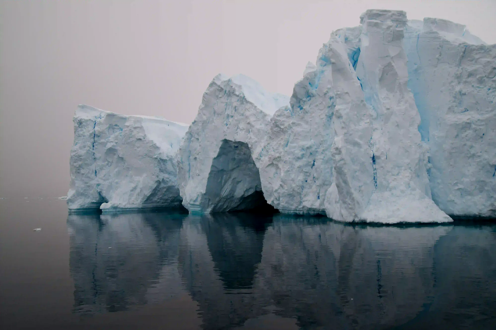 Massive ancient iceberg floating in still, dark water under a muted gray sky, representing the shifting mass of Earth's glaciers.