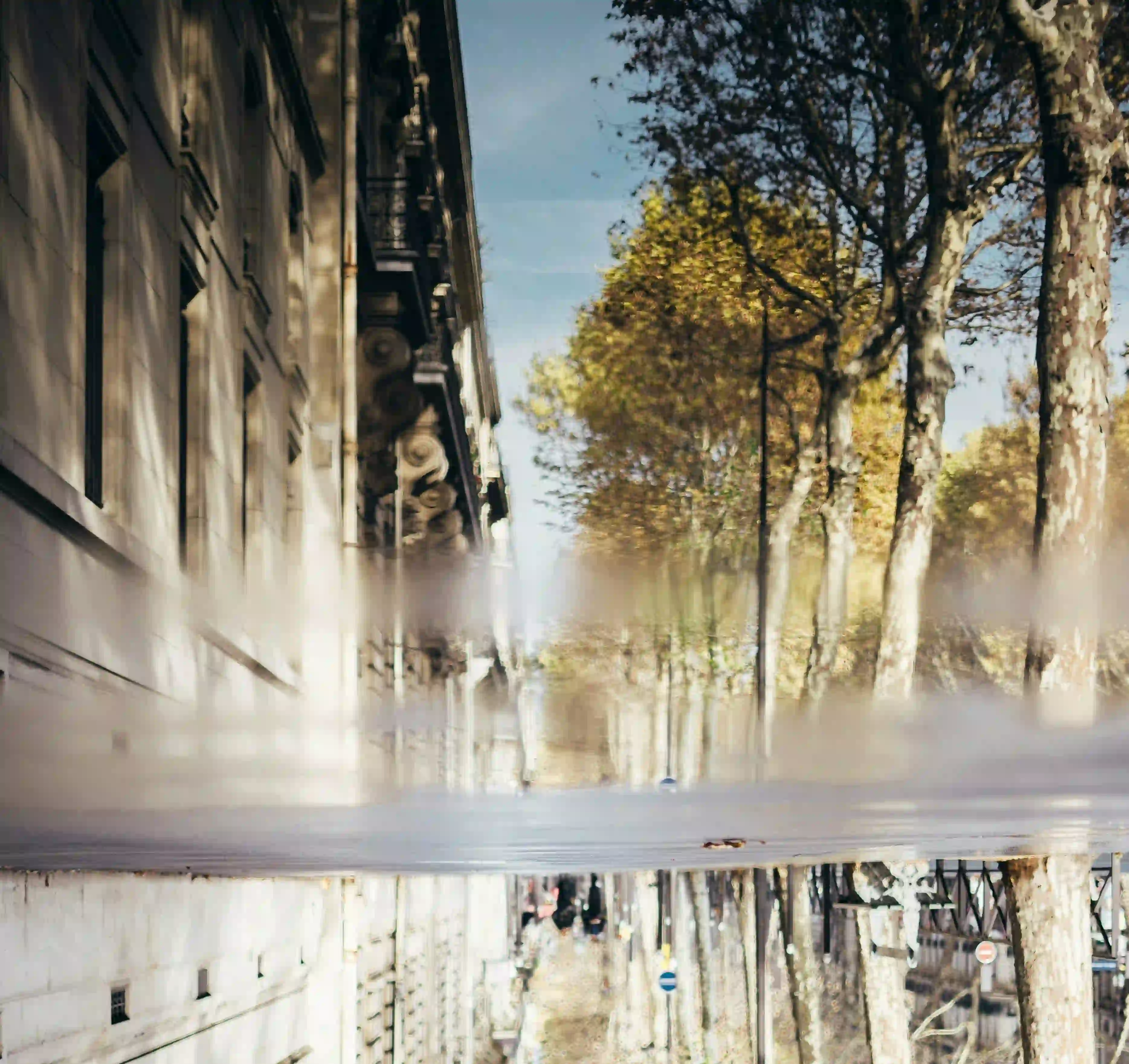 An upside-down reflection of a classic Parisian street with historic buildings and trees, viewed through a puddle on the ground.
