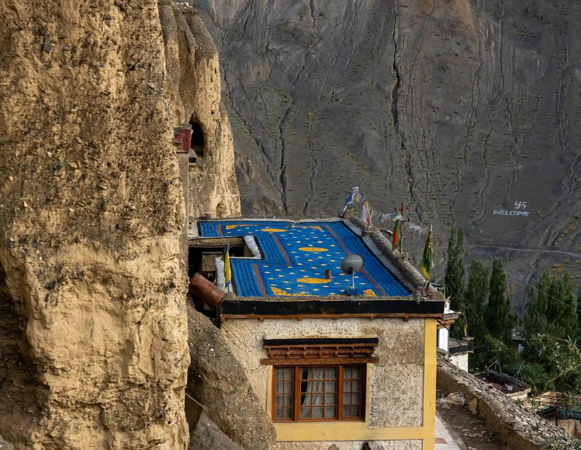 A vibrant blue rooftop art installation with yellow motifs situated on an old stone building built into a cliff face in Lamayuru.