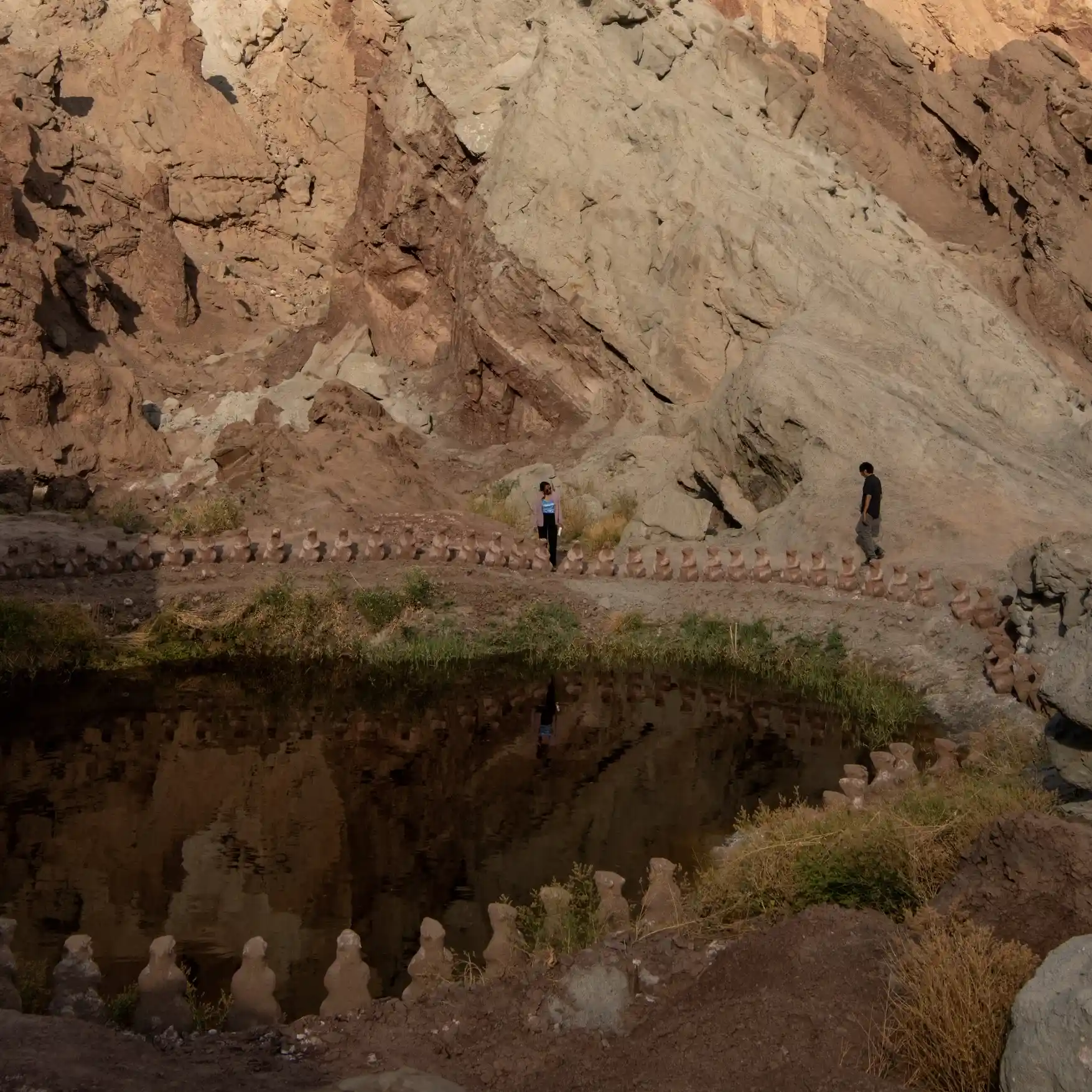A vast, textured rock formation in Basgo towering over a dark pond, bordered by a long line of small clay figures and two tiny human silhouettes.