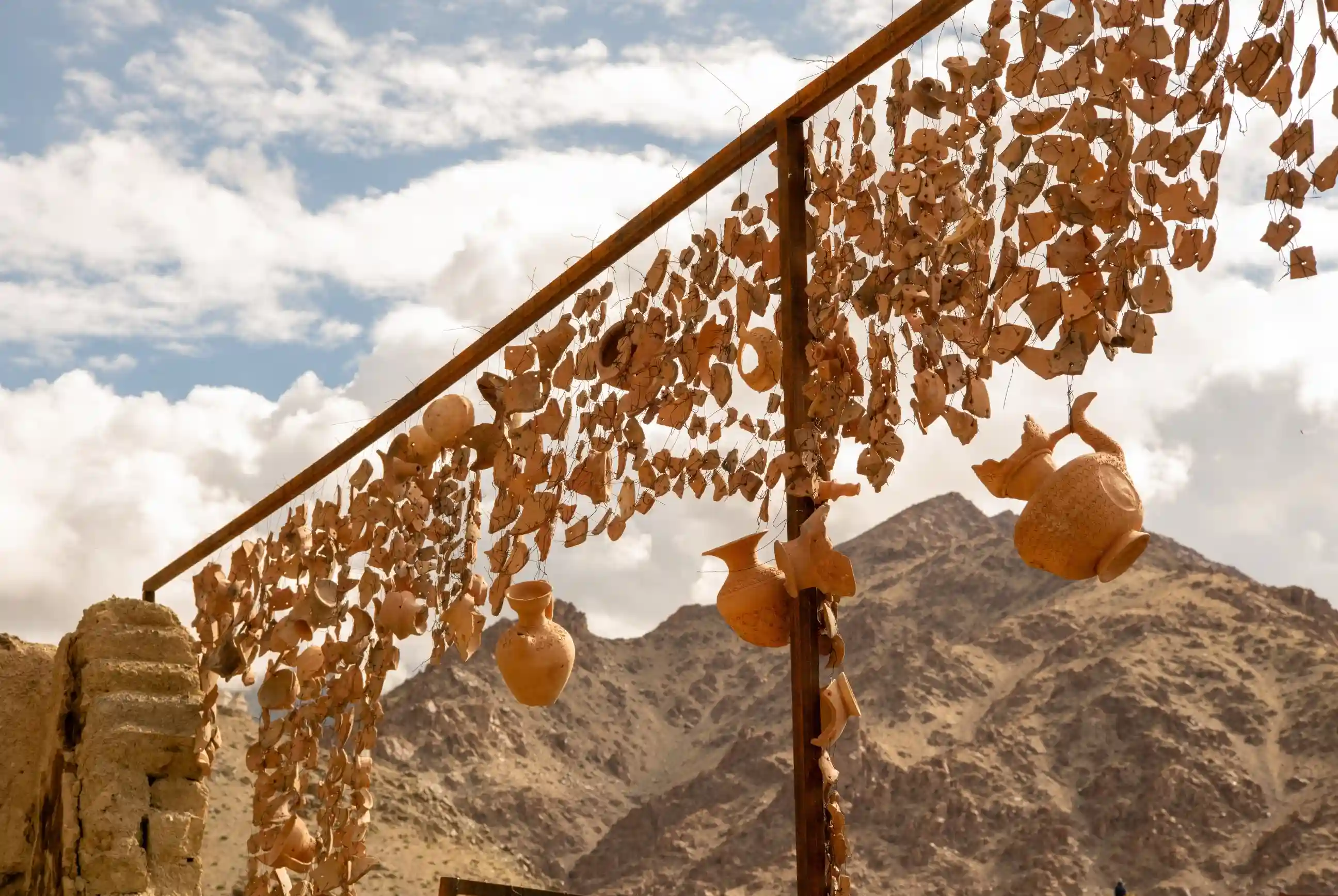 Shattered and whole clay pots suspended from a rusty metal frame against a backdrop of rocky mountains and a cloudy sky in Likir.