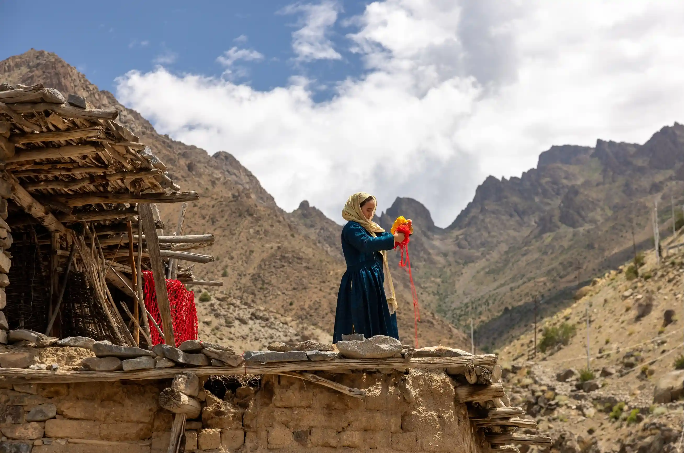 A local woman in traditional blue dress working with vibrant red and yellow threads on a rustic wooden rooftop against a backdrop of jagged Himalayan mountains.