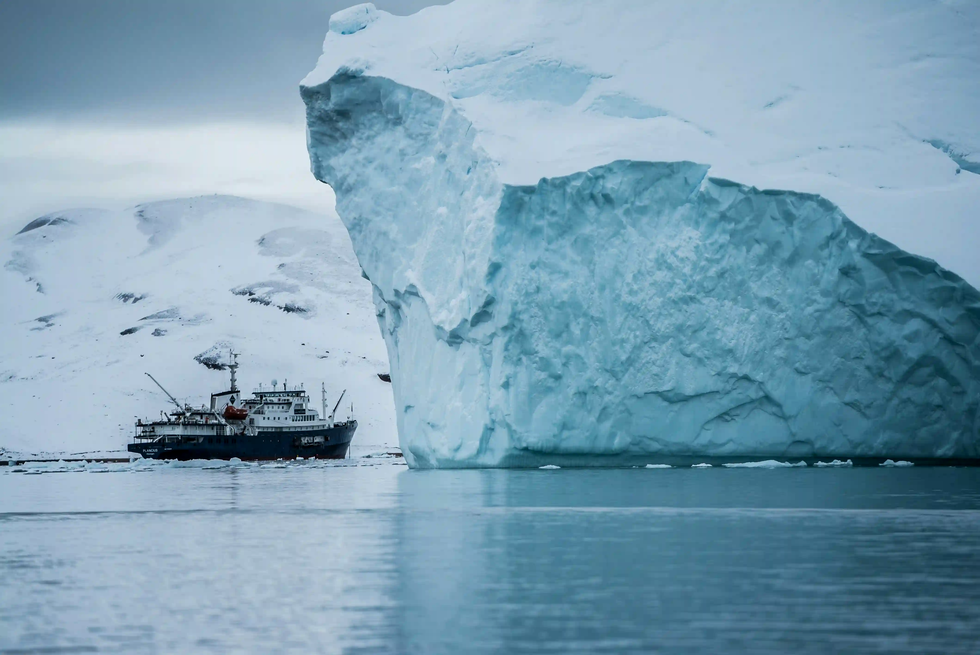 A small ship dwarfed by a colossal blue-white iceberg in a quiet, snowy Arctic fjord under an overcast sky.