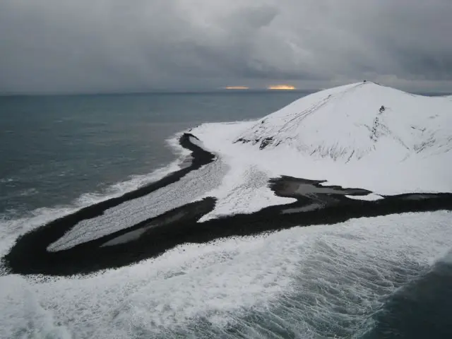 A stark winter aerial view of Surtsey island's north spit covered in snow, showing dark volcanic sand and ocean waves eroding the coastline.