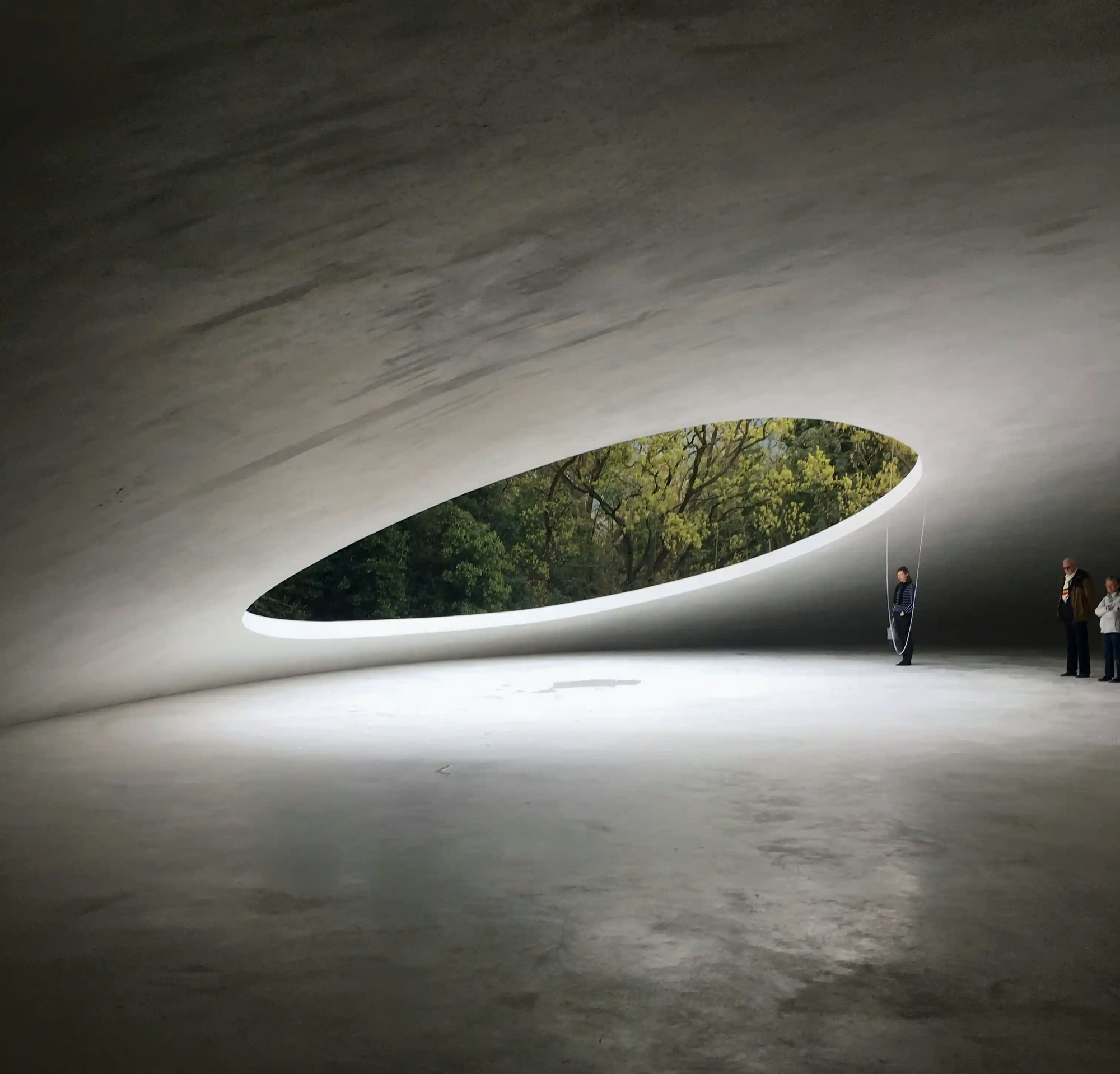 Minimalist curved concrete interior of Teshima Art Museum with a large oval opening revealing green trees and natural light.