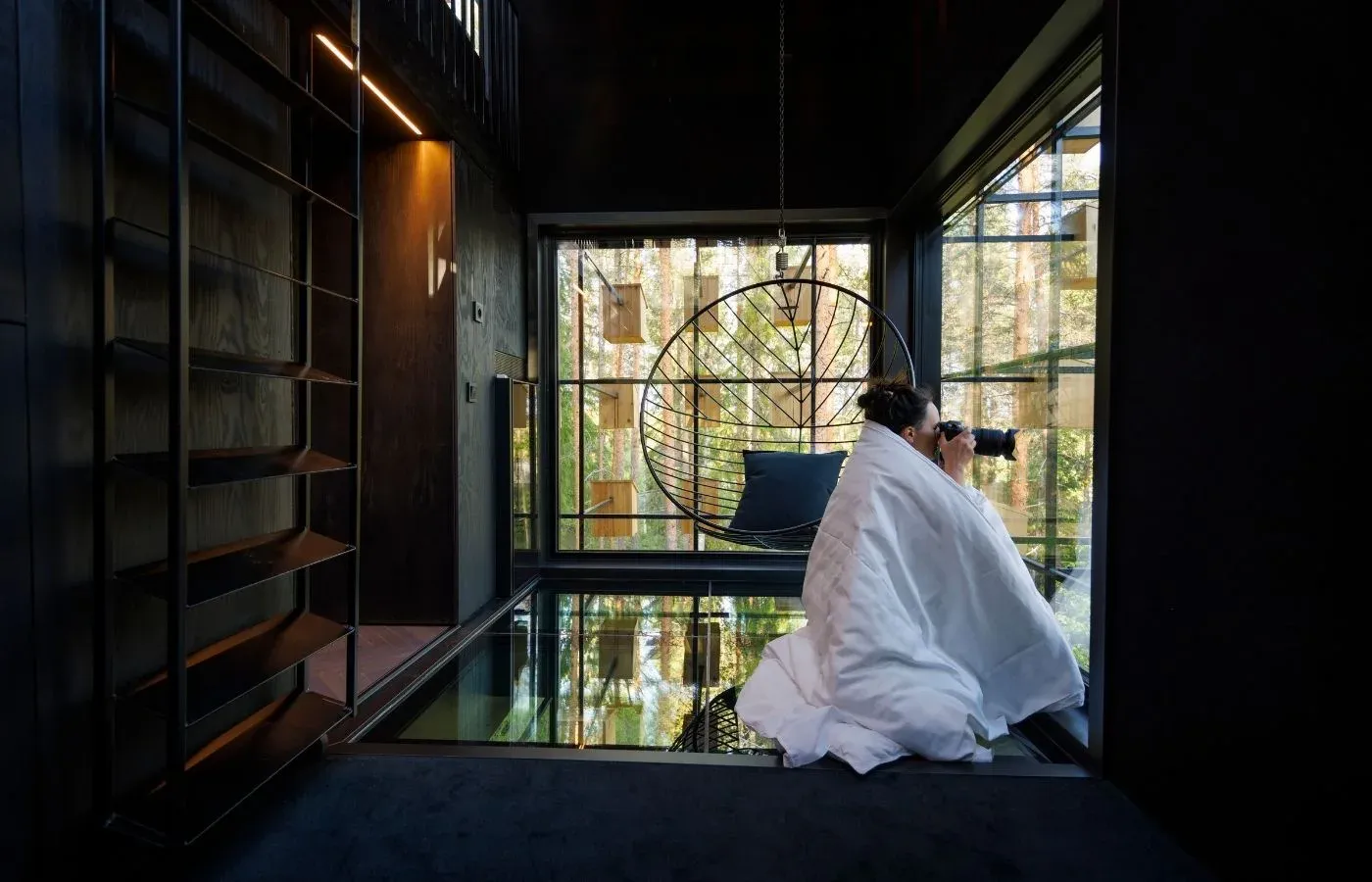 A guest wrapped in a white blanket observes the forest from inside the dark interior of the Biosphere treehouse.