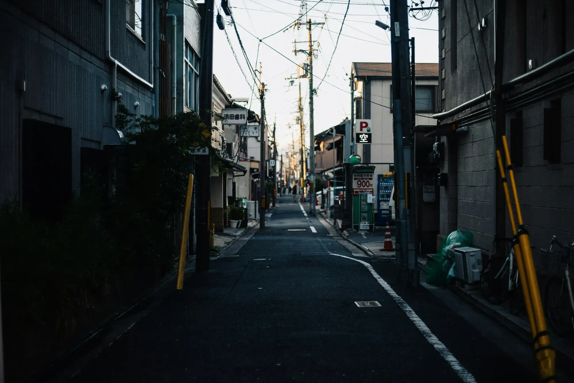 Gloomy, narrow Japanese alleyway flanked by buildings and utility poles, reflecting the urban stress and industrial pressure of the 1980s.