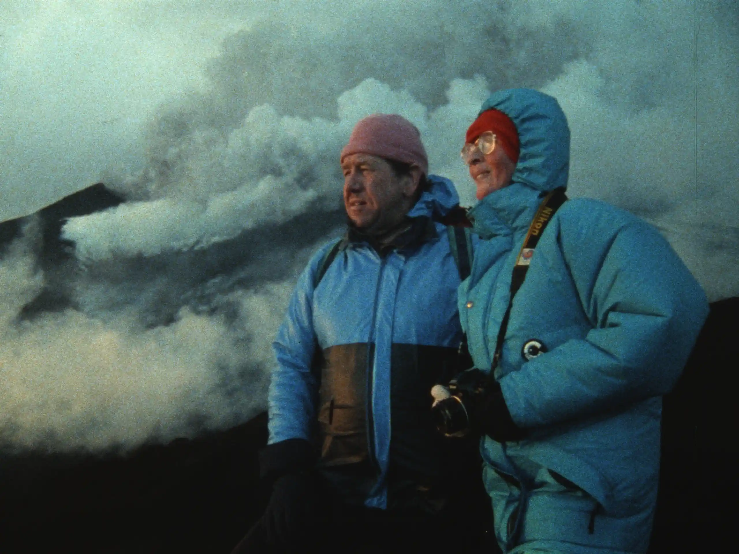 Volcanologists Maurice and Katia Krafft wearing blue winter parkas standing in front of a massive grey ash cloud.