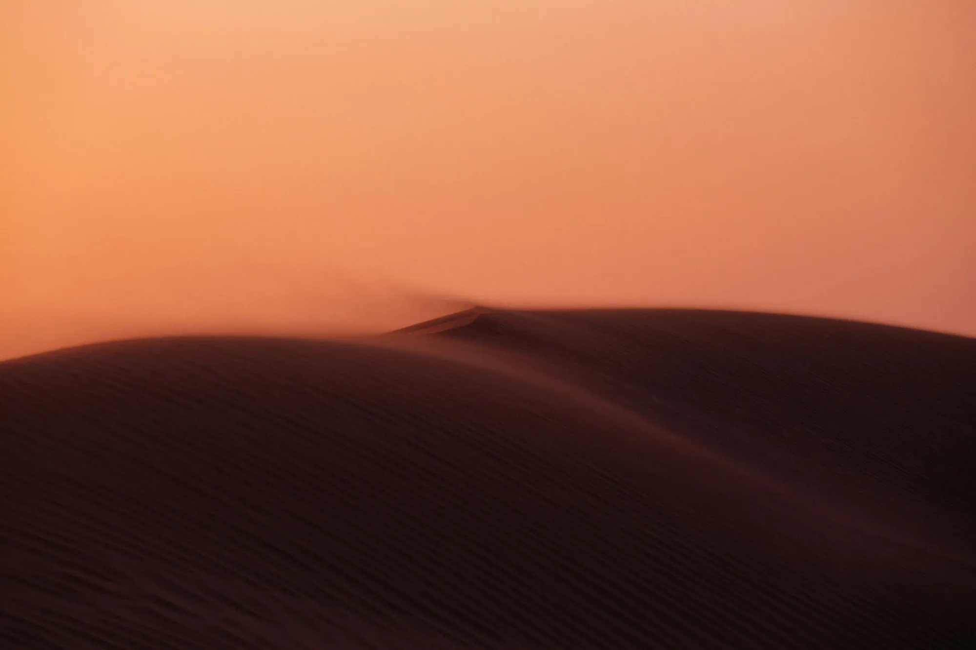 A dark, minimalist view of wind blowing sand over a dune at dusk, representing the concept of invisibility in modern luxury travel.
