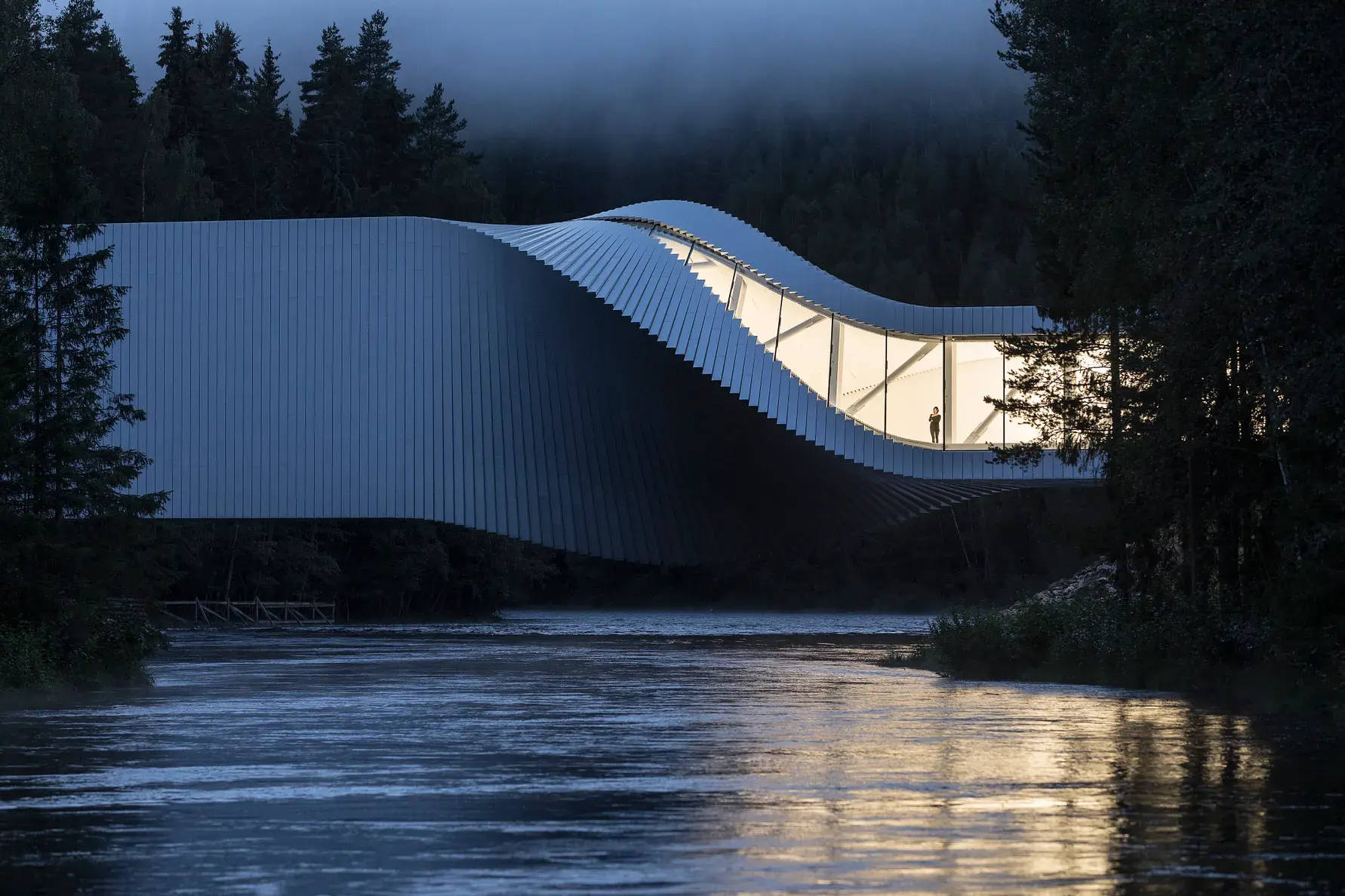 The glowing exterior of The Twist gallery suspended over a river at dusk, showing its 90-degree structural contortion and a person standing inside the glass walkway.