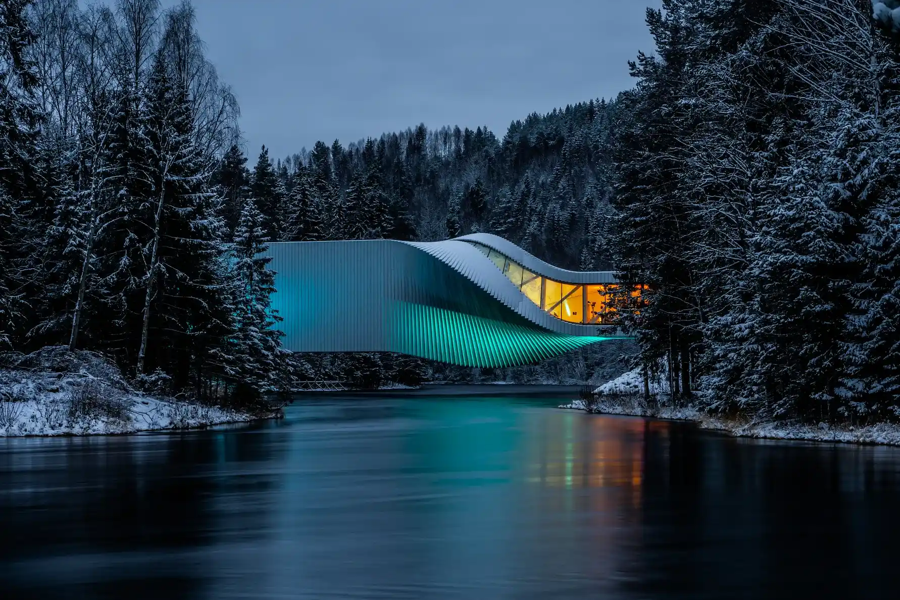 The Twist gallery illuminated with teal and warm lights, framed by snow-covered pine trees and a dark river during a winter evening.