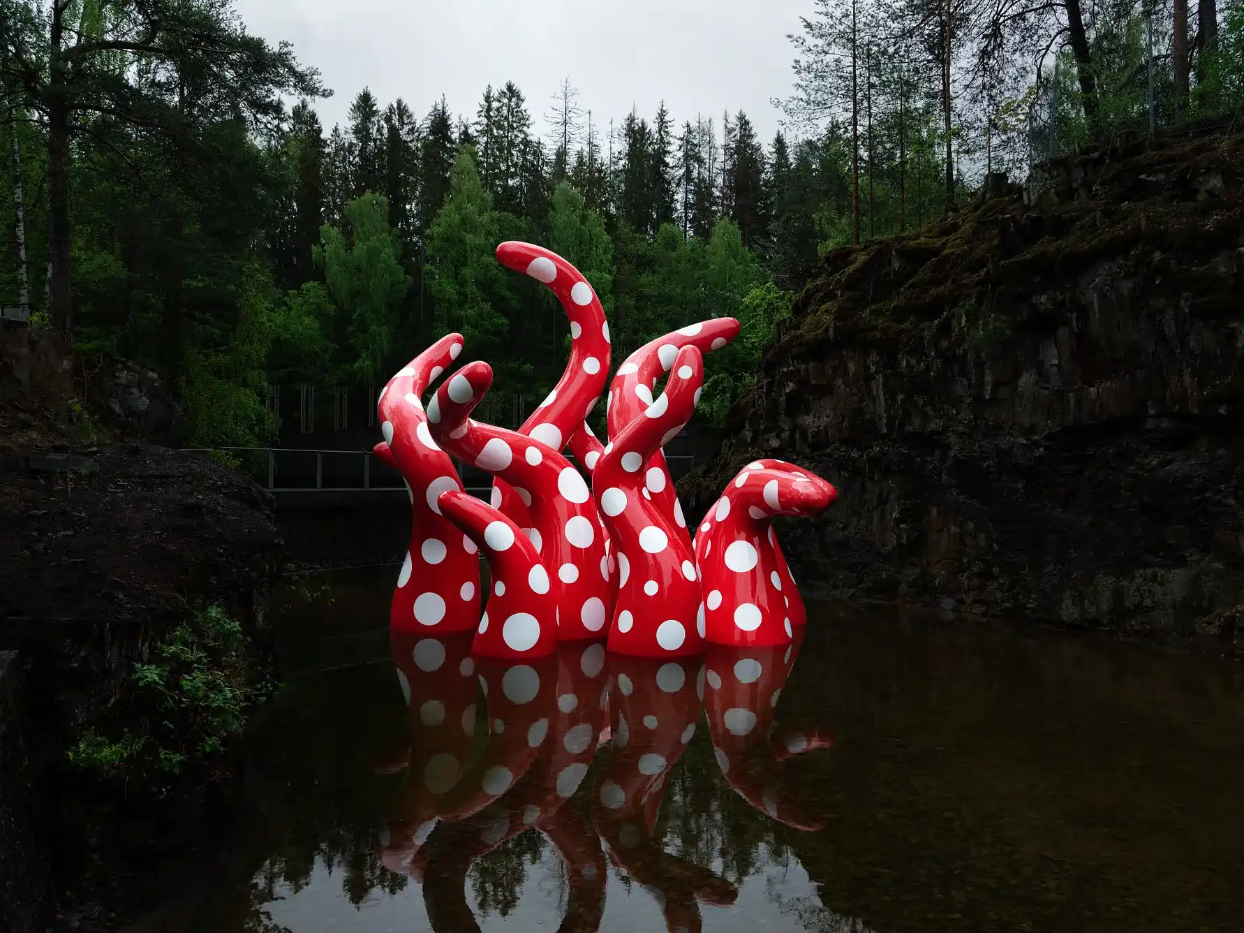 Red and white polka dot tentacles of Yayoi Kusama's Shine of Life sculpture emerging from the dark waters of a river surrounded by a Norwegian forest.