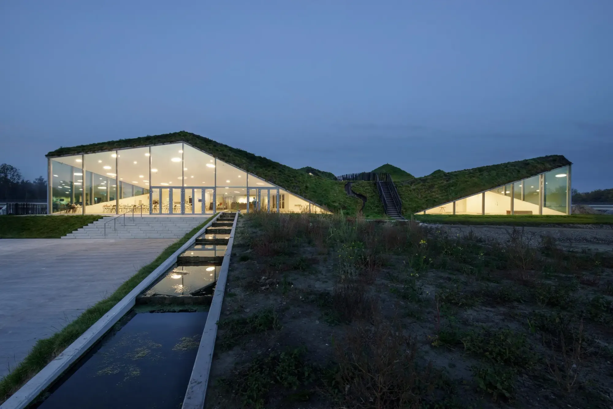 A modern museum building with a large sloping green grass roof flanked by water channels at dusk.