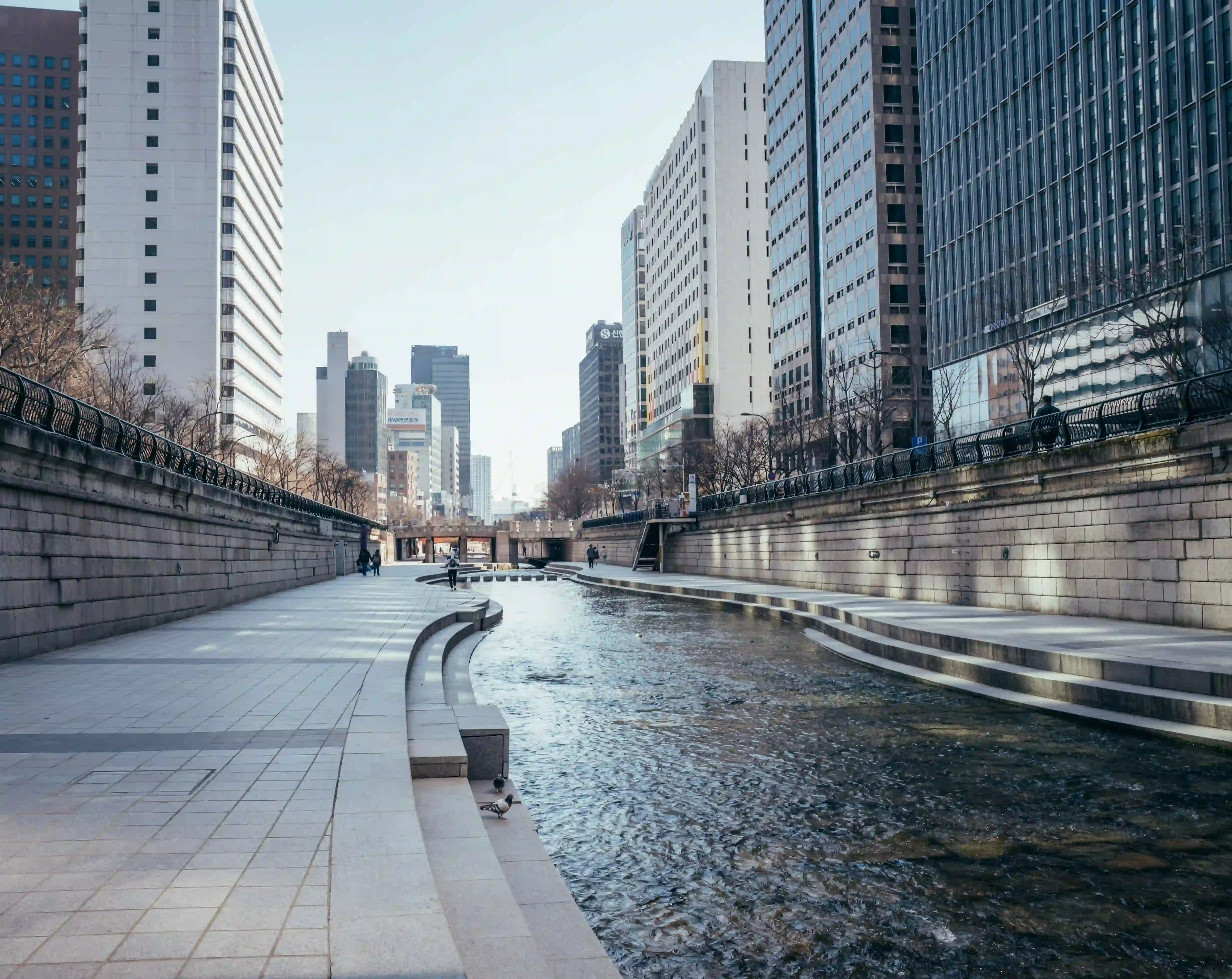 A cold and structured view of the Cheonggyecheon stream in Seoul, heavily bordered by sharp concrete infrastructure and towering high-rises.
