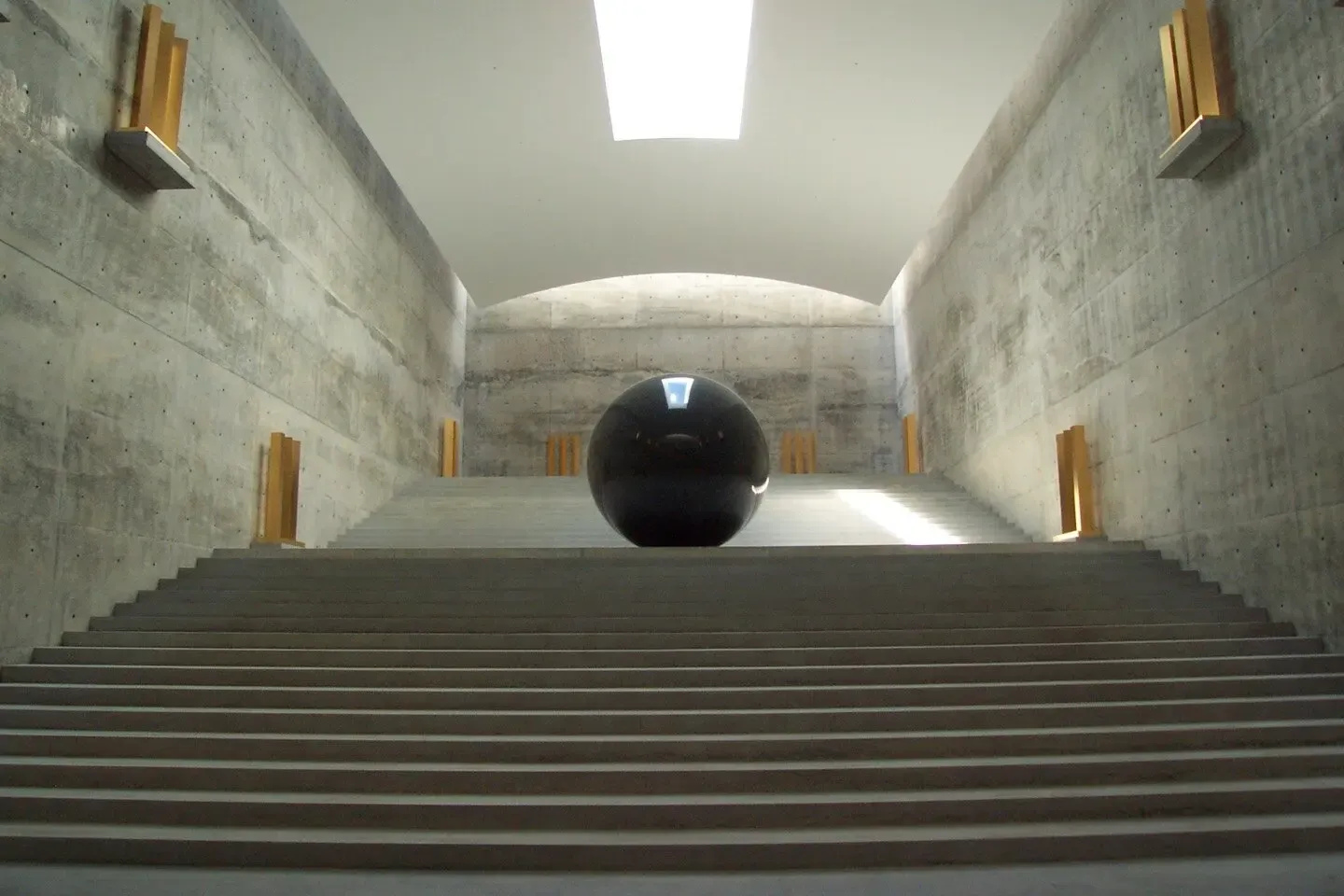 A large smooth black sphere rests on concrete stairs under a geometric skylight inside the underground Chichu Art Museum.
