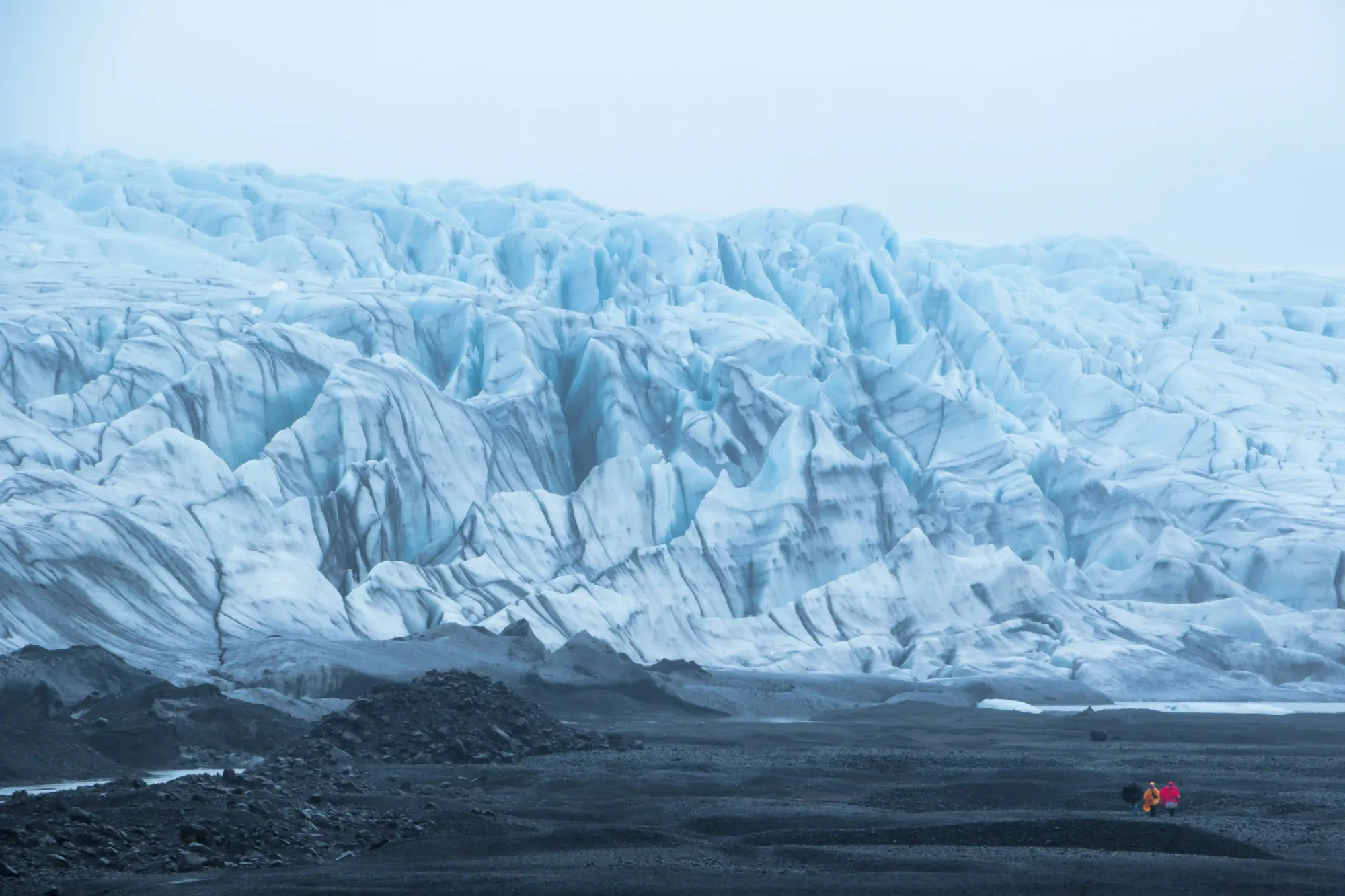 A vast, rugged icy glacier under an overcast sky, with tiny human figures visible in the dark foreground.