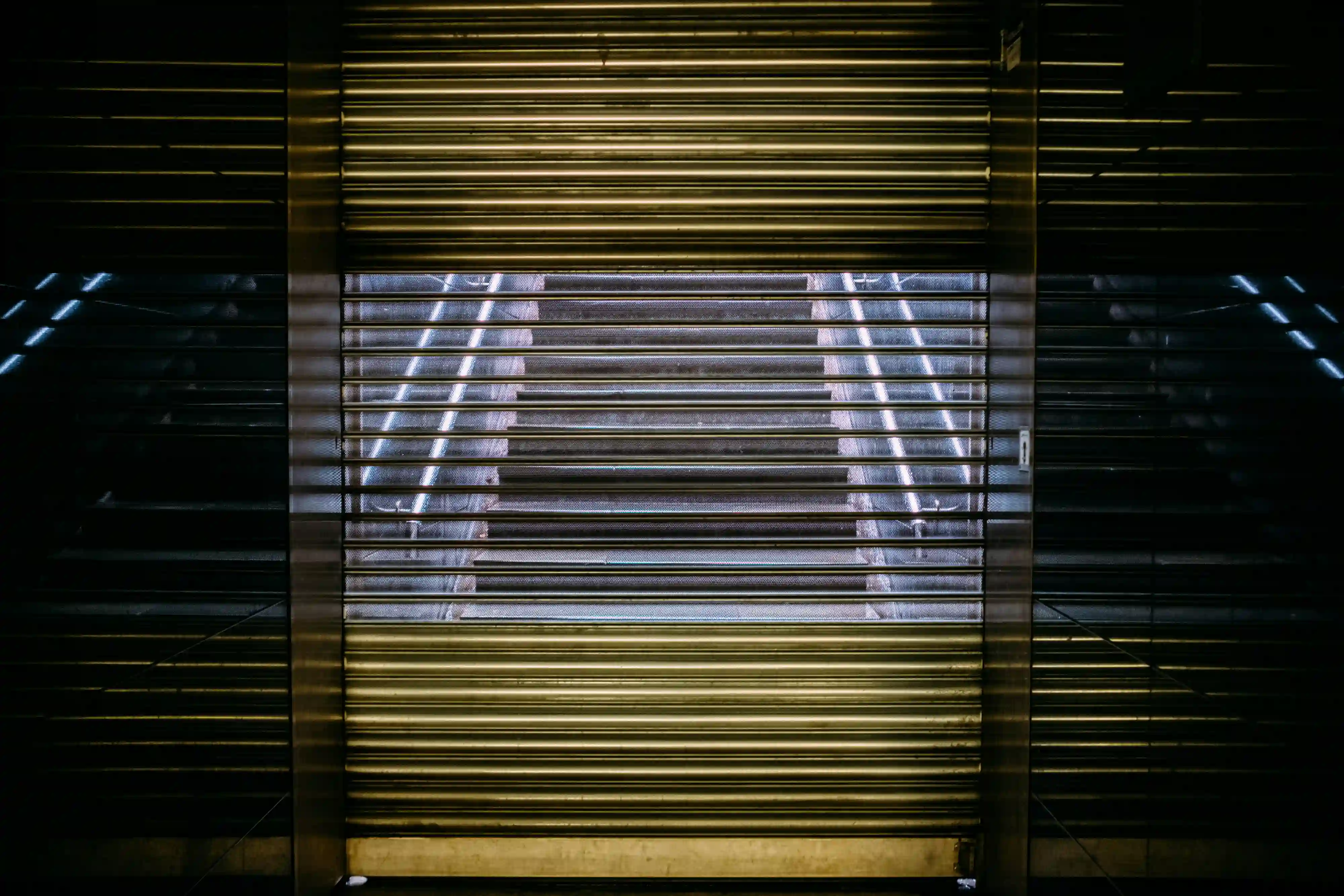 A dark, moody view through a partially open industrial metal shutter revealing illuminated stairs, symbolizing the restricted physical reality of data centers.