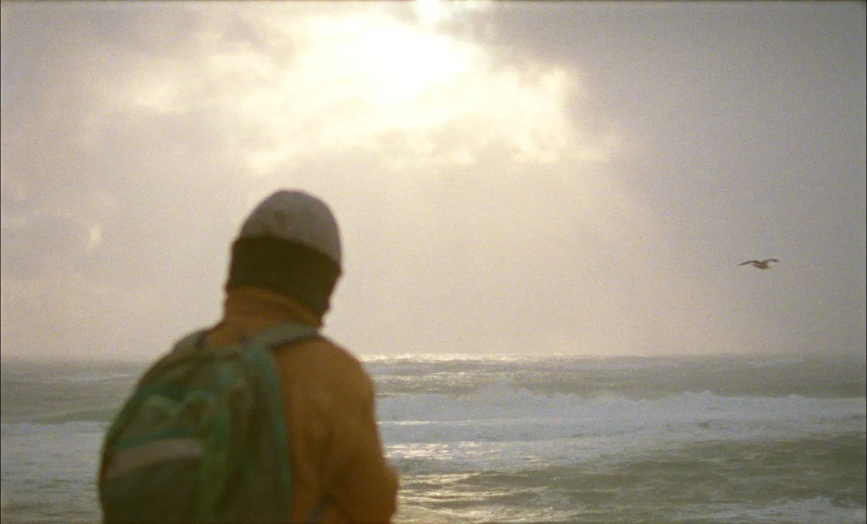 16mm film still of Zoe Lucas looking out at the ocean on Sable Island with sunlight breaking through overcast skies in Geographies of Solitude