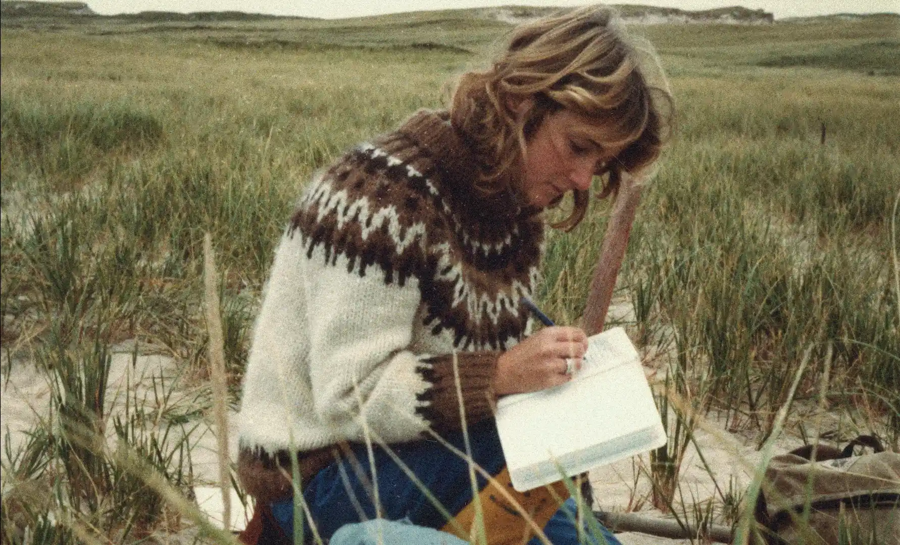 Archival slide photo of a young Zoe Lucas working as a researcher, writing in a field notebook outdoors on Sable Island in Geographies of Solitude.