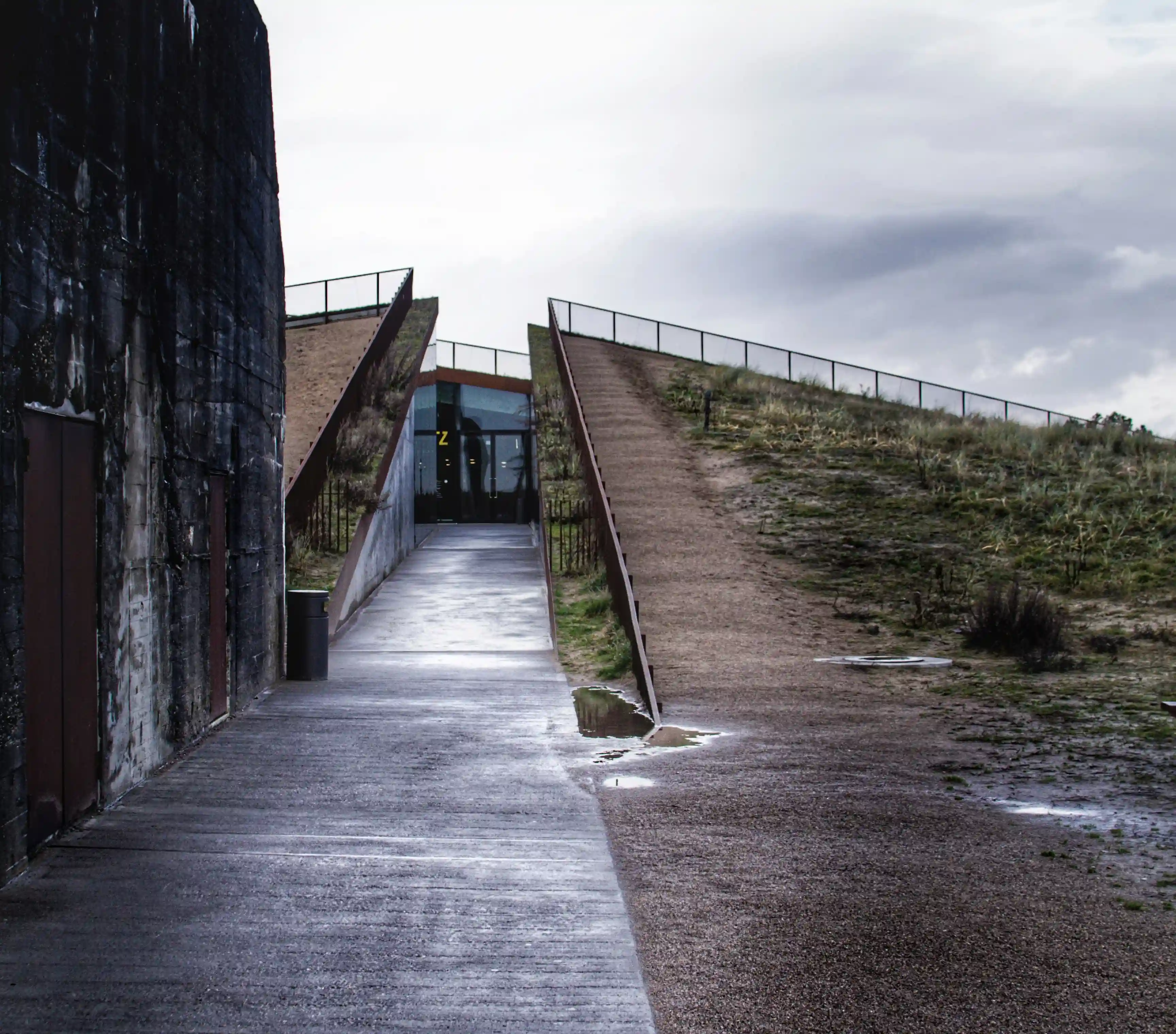 Concrete pathways slicing through grassy dunes leading to the glass entrance of the Tirpitz Museum, next to a dark World War II bunker.