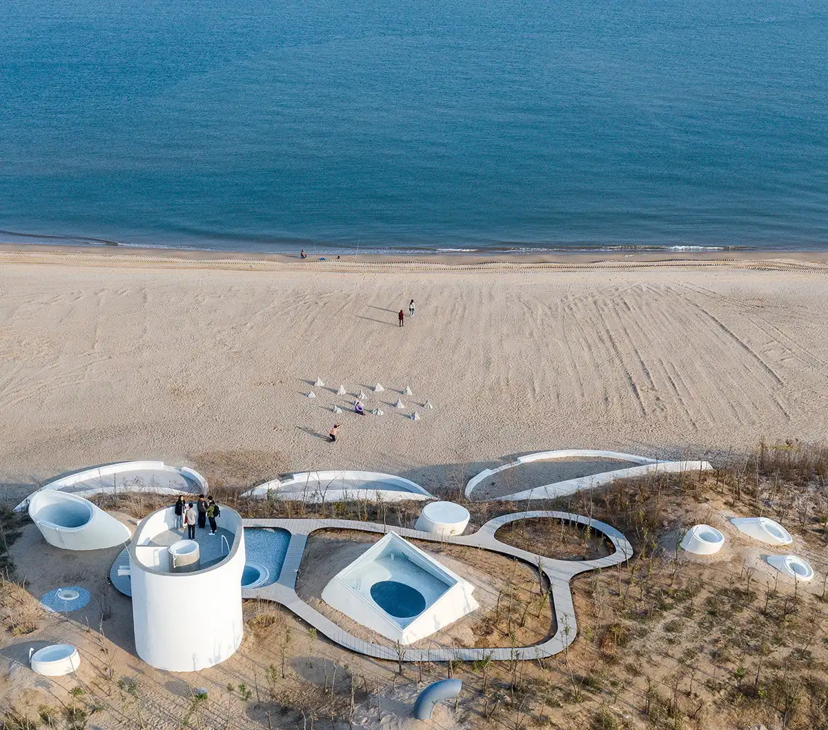 Aerial view of the UCCA Dune Art Museum with white organic concrete forms partially buried in the sand on a beach in China.