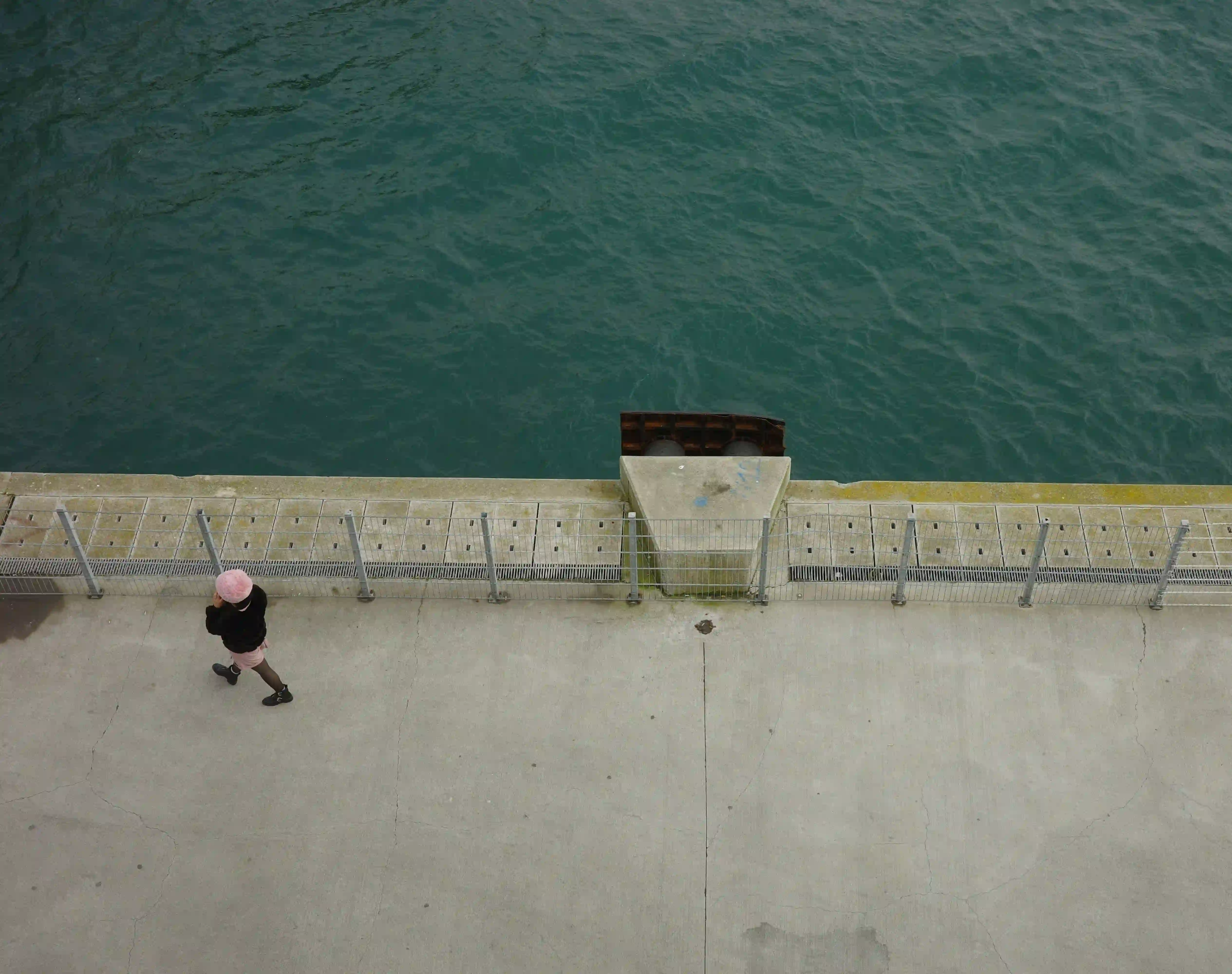 A high-angle, desaturated shot of a solitary person walking along a heavily fenced concrete waterfront, capturing the alienation of urban gentrification.