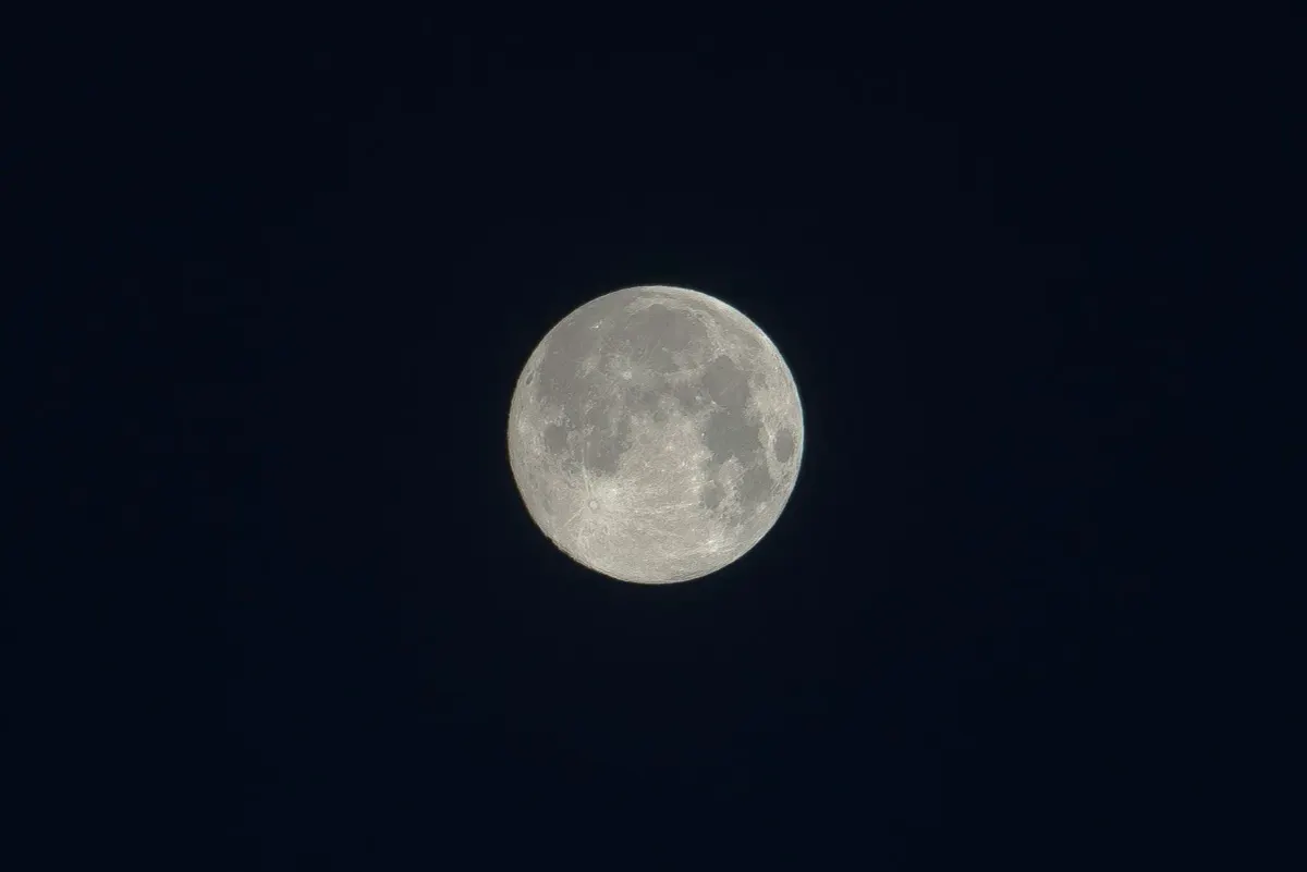 A highly detailed, pale full moon glowing against a pitch-black night sky, revealing its true color rather than a blue hue.