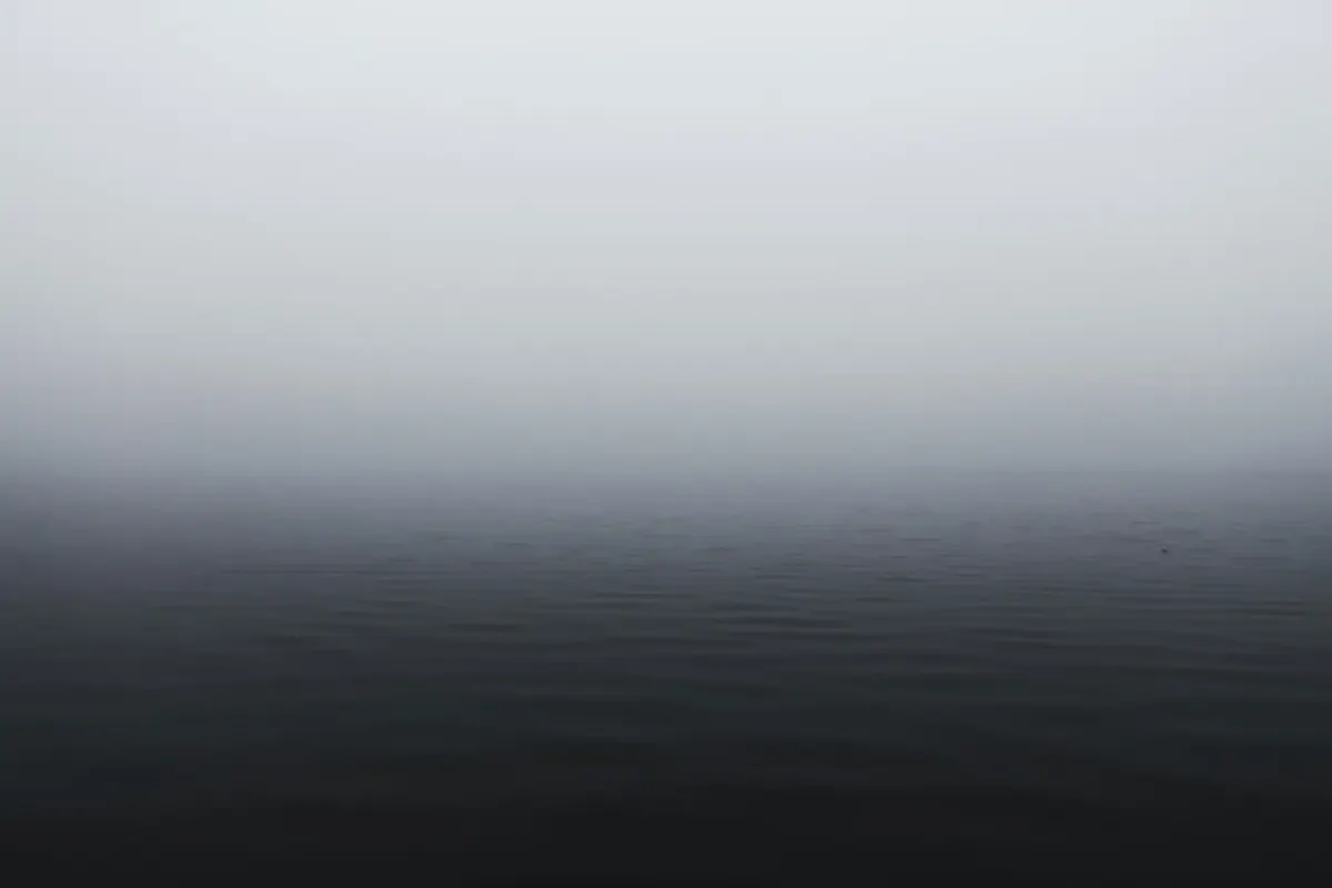 A minimalist and moody view of a calm, dark body of water merging completely into a dense, gray fog, obscuring the horizon.