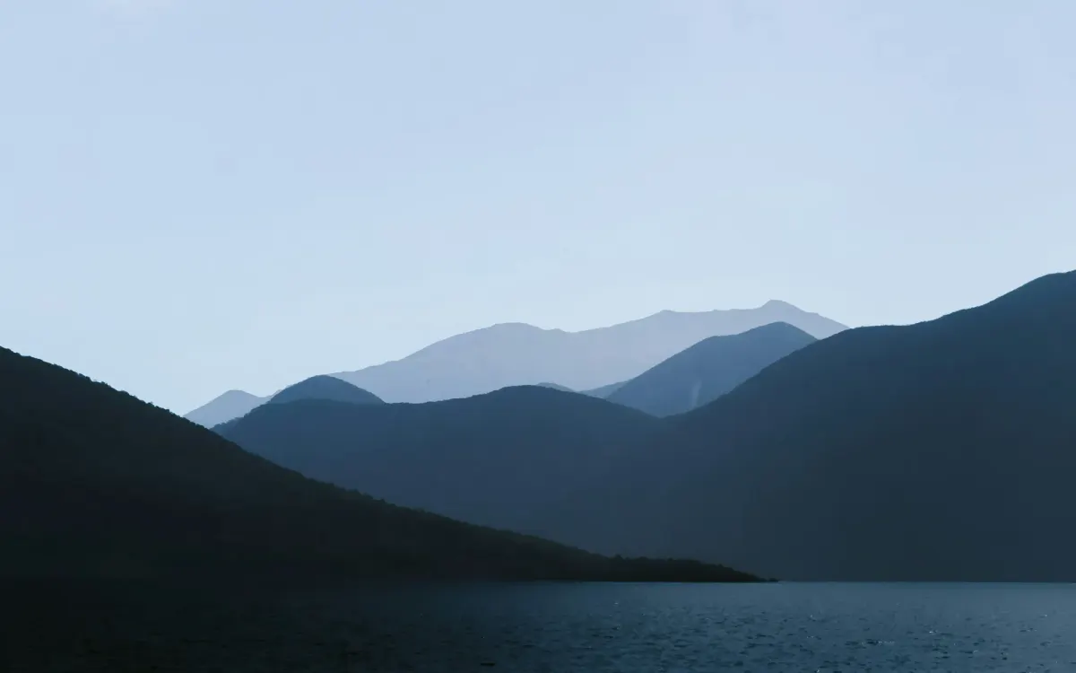 A serene landscape featuring layered, dark mountain silhouettes fading into a pale, misty sky above a calm body of water.