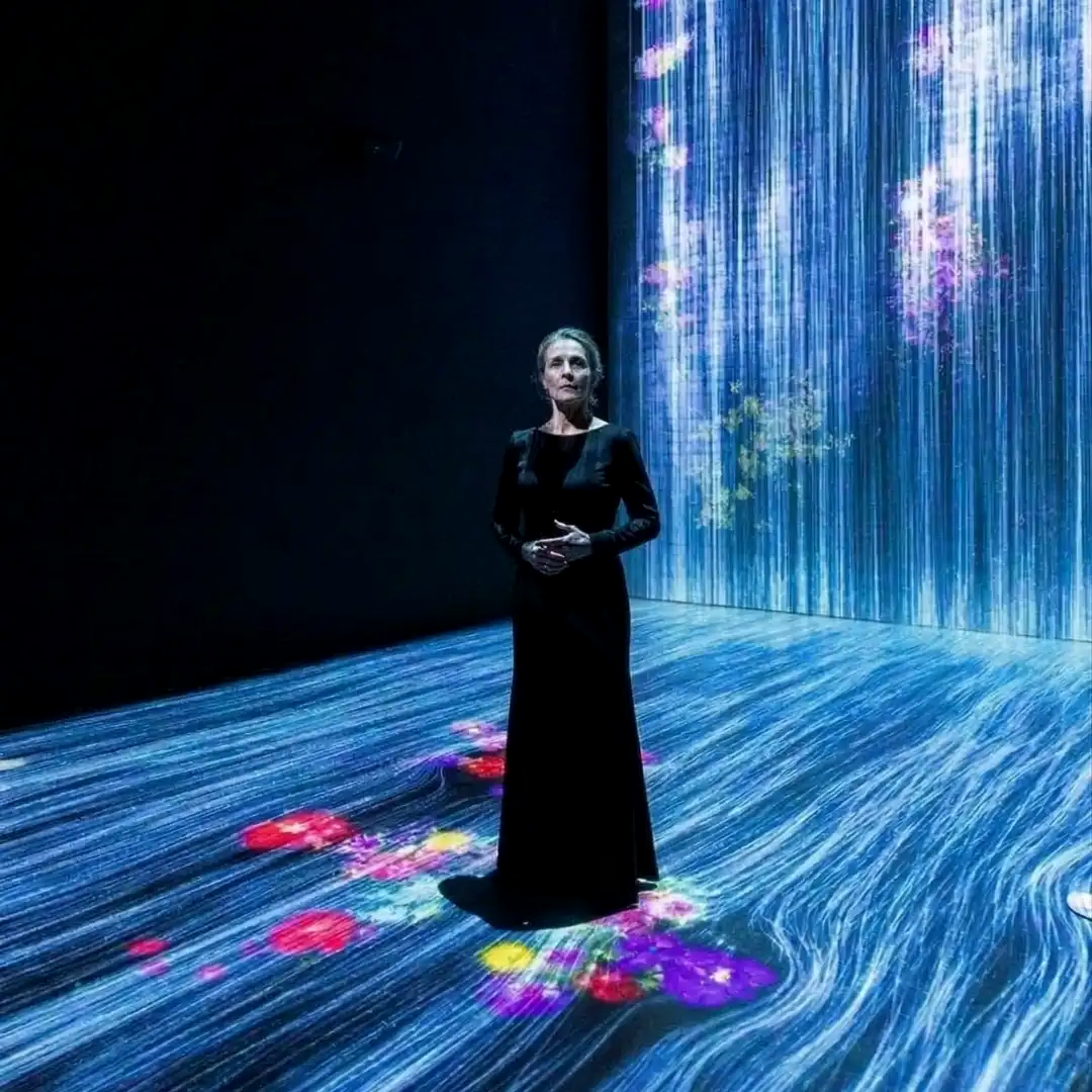 A visitor standing alone in a dark, immersive museum exhibition illuminated by digital projections of flowing water and glowing flowers.