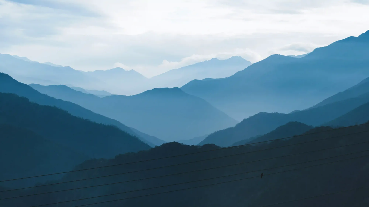 Layered blue mountain ridges fading into thick mist under an overcast sky, with subtle power lines in the dark foreground.