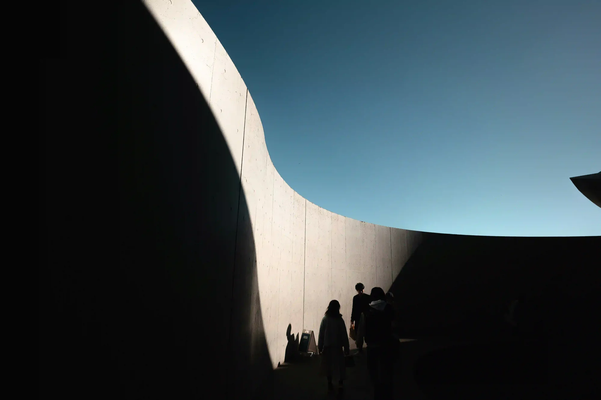 Silhouettes of people walking along a massive curved concrete wall divided by deep shadows, representing urban isolation and architectural silence.