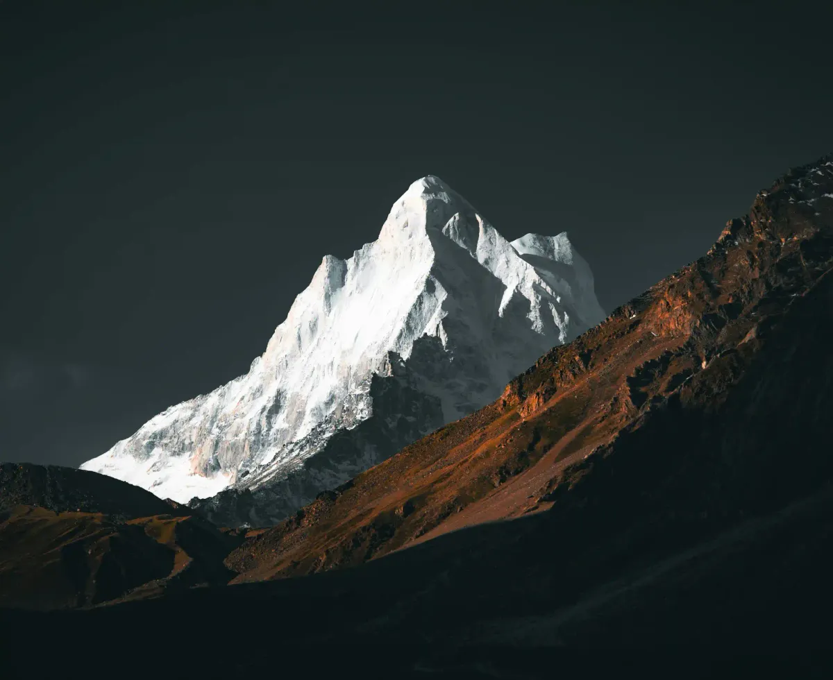 A dramatic, sunlit snow-capped mountain peak emerging from deep shadows, showcasing the stark and harsh geology of the planet.
