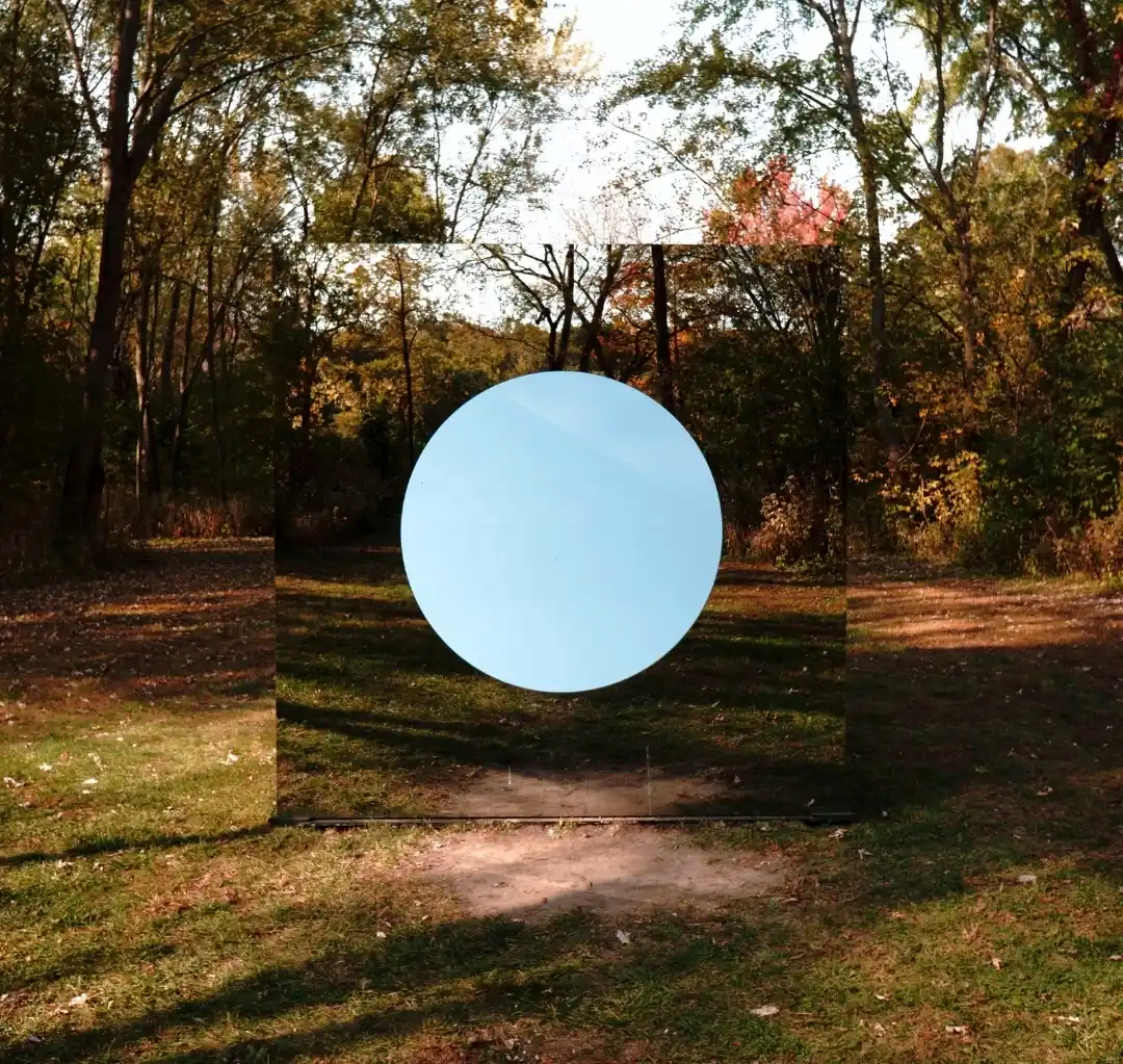 A large square mirror reflecting a forest stands in the middle of the woods, featuring a solid pale blue circle in its center, creating a surreal visual illusion.