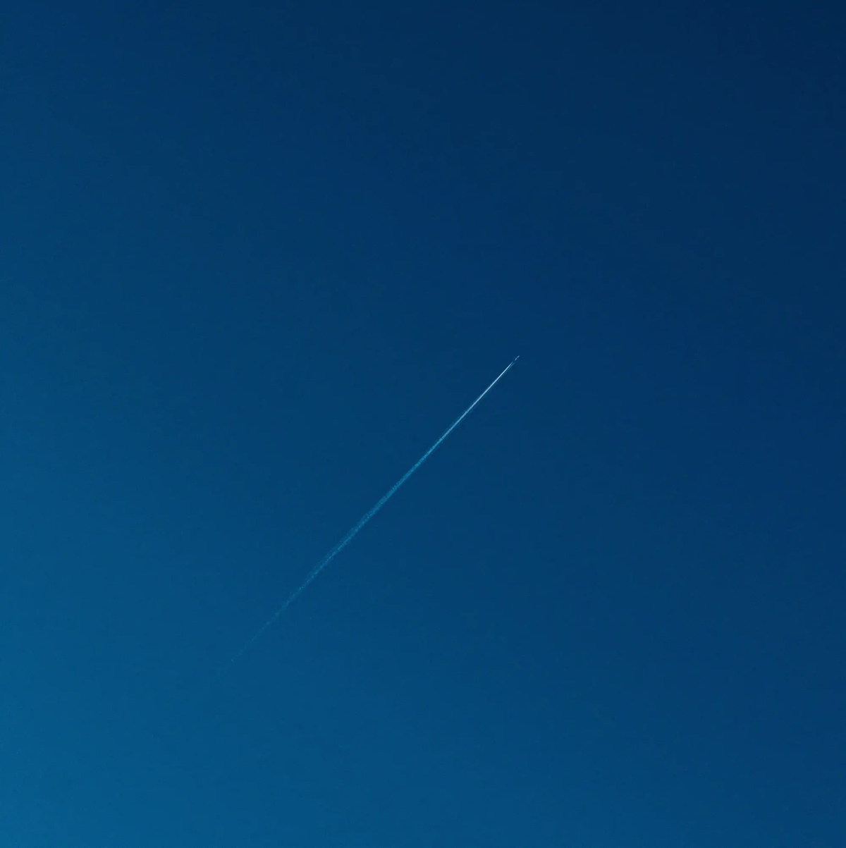 A single airplane leaving a thin white contrail across a vast, deep blue minimal sky.
