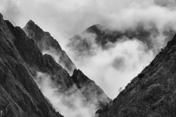 A dramatic black-and-white landscape of rugged, steep Andean mountain peaks partially swallowed by dense, swirling mist in the Peruvian cloud forest.