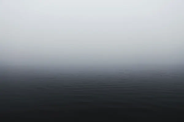 A minimalist and moody view of a calm, dark body of water merging completely into a dense, gray fog, obscuring the horizon.