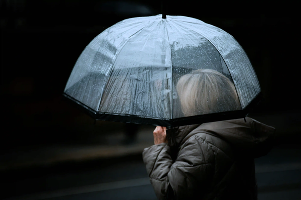 A person seen from behind holding a clear umbrella covered in raindrops against a dark, blurred urban background.