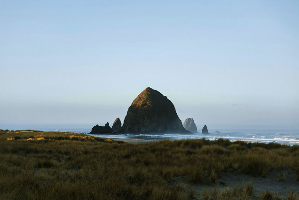 A serene view of Haystack Rock at Cannon Beach, Oregon, featuring golden coastal grass and misty ocean waves for a cool northern summer escape.