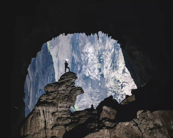 A tiny silhouette of a person standing on an underground rock formation, highlighting the colossal scale and natural light of a massive cave system.