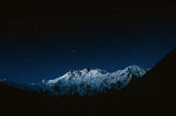A vast, dark night sky filled with faint stars above a snow-capped mountain silhouette, representing the silent scale of the universe.