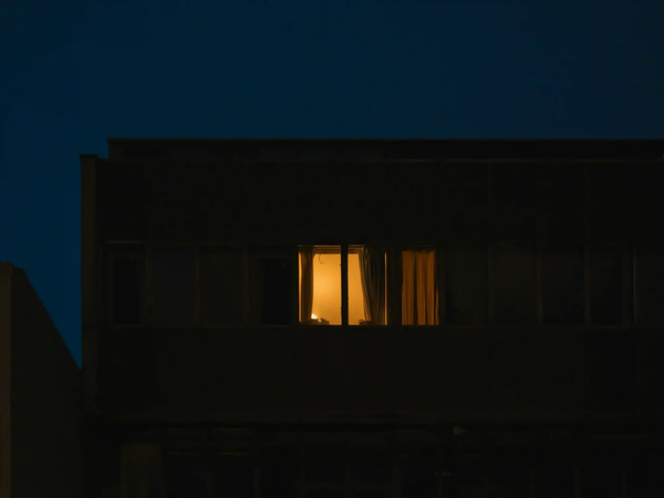 A single illuminated window in a dark, imposing concrete building at night, representing the feeling of urban isolation and solastalgia.