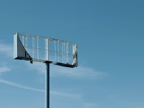 A skeletal, empty billboard frame with torn pieces of canvas hanging against a clear sky, symbolizing the removal of outdoor advertising and the stark reality of visual silence.