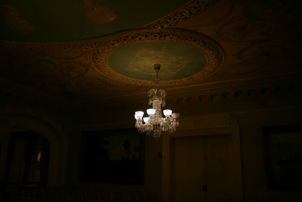 A glowing crystal chandelier illuminating the ornate ceiling of a dark, historic room.