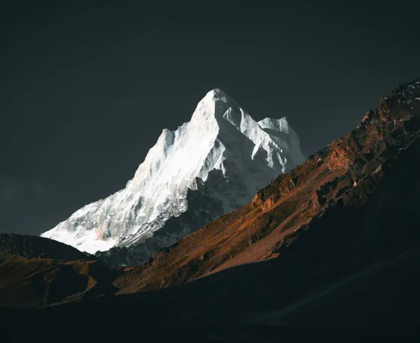 A dramatic, sunlit snow-capped mountain peak emerging from deep shadows, showcasing the stark and harsh geology of the planet.