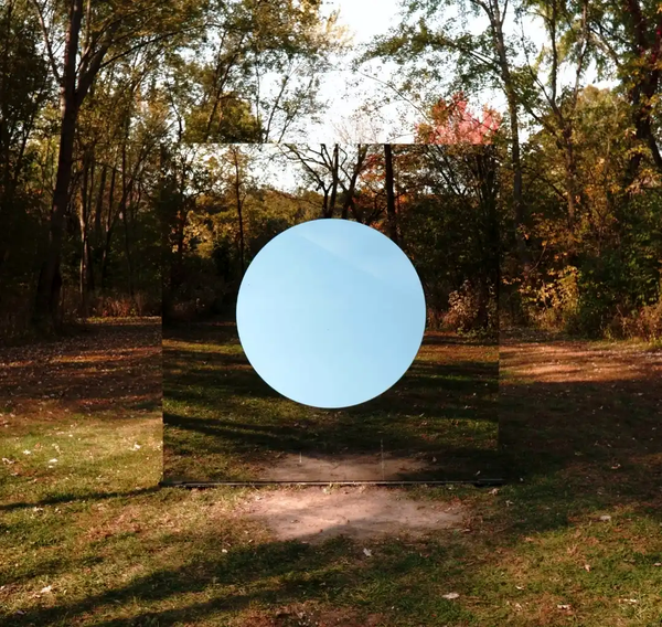 A large square mirror reflecting a forest stands in the middle of the woods, featuring a solid pale blue circle in its center, creating a surreal visual illusion.
